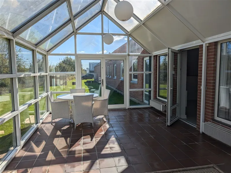 Terrasse med glasvinduer, hvide stole og et rundt bord placeret midt på terrassen. På loftet hænger to hvide kuglelys.