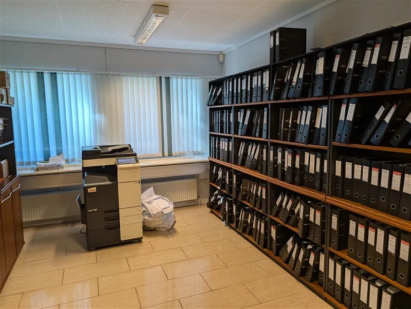 Kontor med et printer og et bibliotek med mange sorte mapper og papirkurve samt radiator ved vinduet