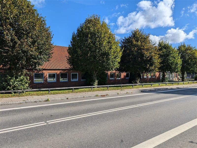 Et lille erhvervsområde med røde mursten og tæt plantet grønt i baggrunden ved siden af en bred asfaltvej