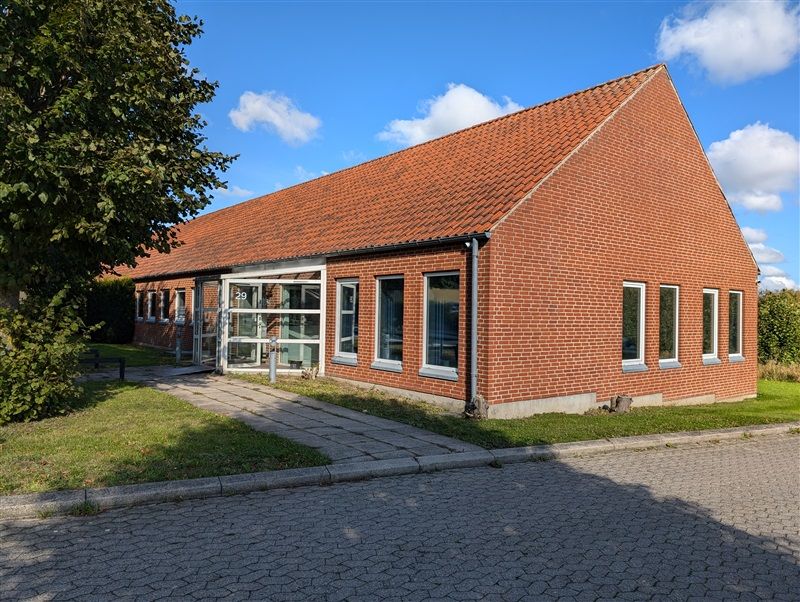 Rød teglstensbygning med rødt tag, vinduer og glasdør, nummer 29, fuld af grønne buske og sneglestensvej