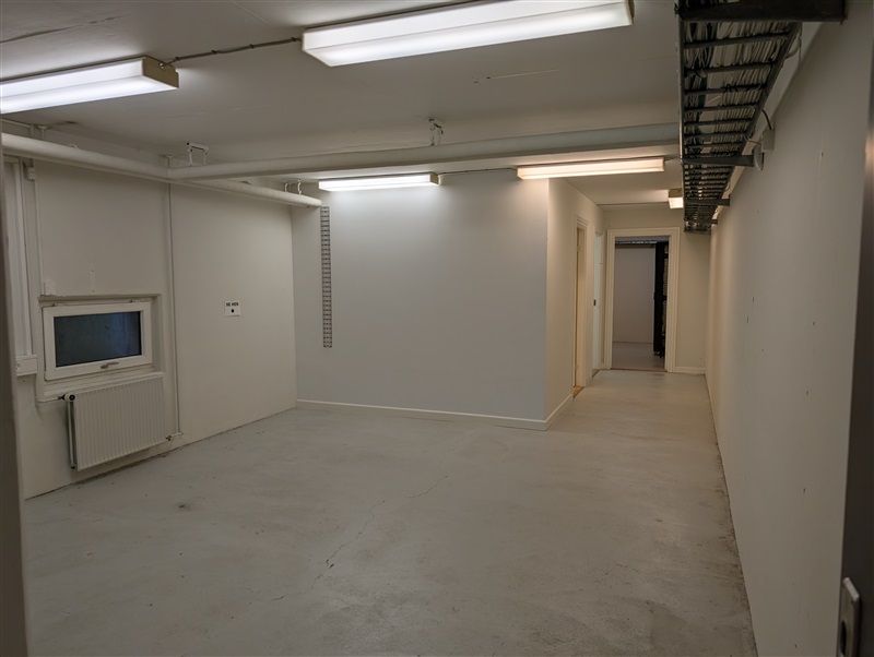 Lang, tom gange med hvide vægge og gulv. På højre side er døren åben, og på venstre side er et vindue med en radiator.