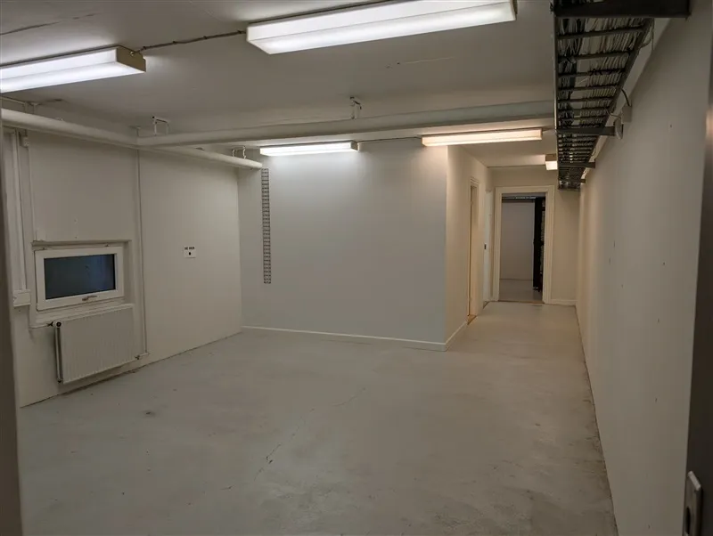 Lang, tom gange med hvide vægge og gulv. På højre side er døren åben, og på venstre side er et vindue med en radiator.