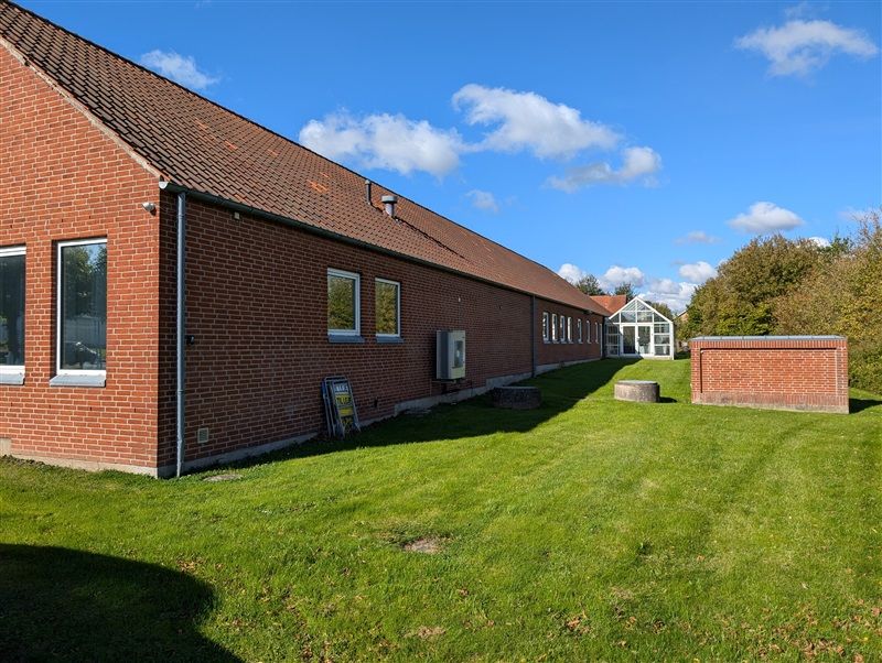 En rødmalet bygning med glasruder og en skur udenfor samt grønt græs og træer i baggrunden