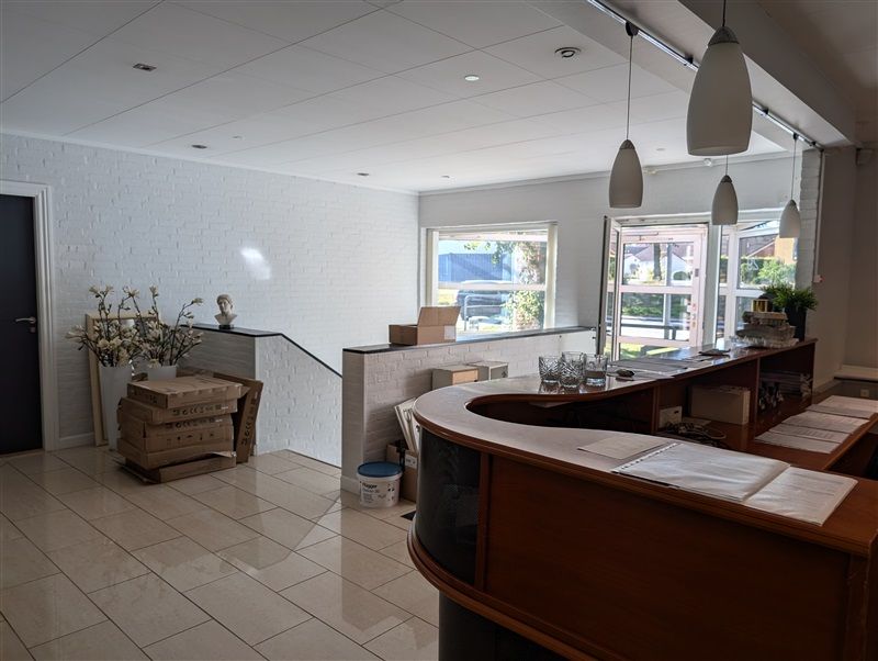 Erhvervsejendom med stort lobbyområde, møbler, kufferter, lamper og blomster på en høj hylde samt vinduer mod en gade