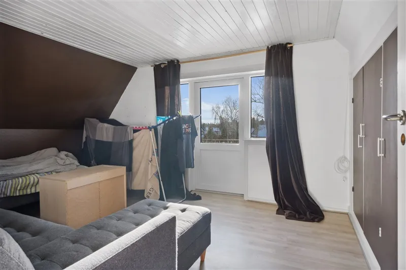 Lille værelse med højloft, hvidt loft og vægge, gulvbelægning, sofa, seng, tøjstativ og to hængekurve med tøj