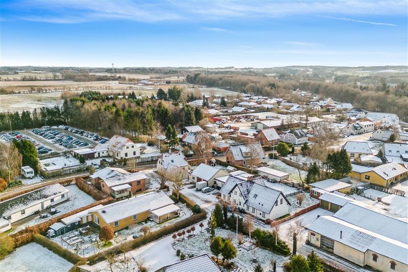 Et landsbybillede med huse og gader dækket af sne og træer, et stort parkeringsområde, og i baggrunden bjerge og marker