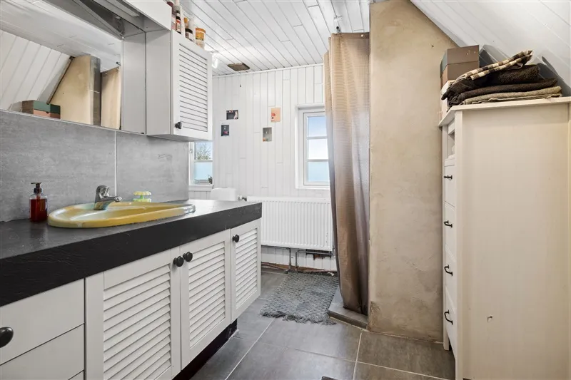 Foto af et lille badeværelse med en hvid væg og loft samt en hvid radiator. Der er en hvid skab med håndklæder samt en