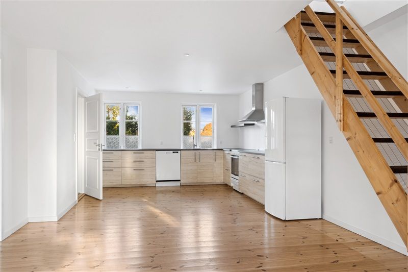 Et moderne og velholdt køkken med hvide vægge, lysegrå loft, trægulv og lysegrå apparater, herunder køleskab, vaskemaskine