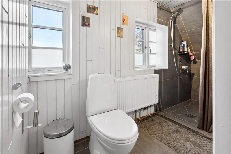 Toiletrum med hvid toilet, radiator, skam, to toiletpapirruller og en lukket garderobe.