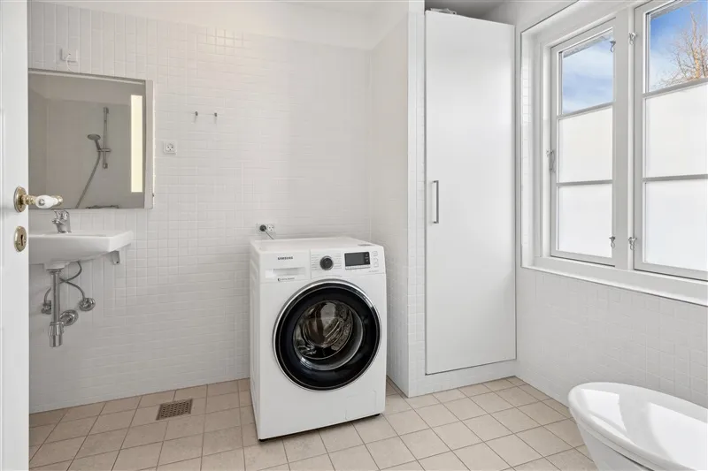 Moderne badeværelse med hvid vasketøjsmaskine, hvidt badekar, hvidt spejl, hvidt håndvask og hvidt badeværelseskab