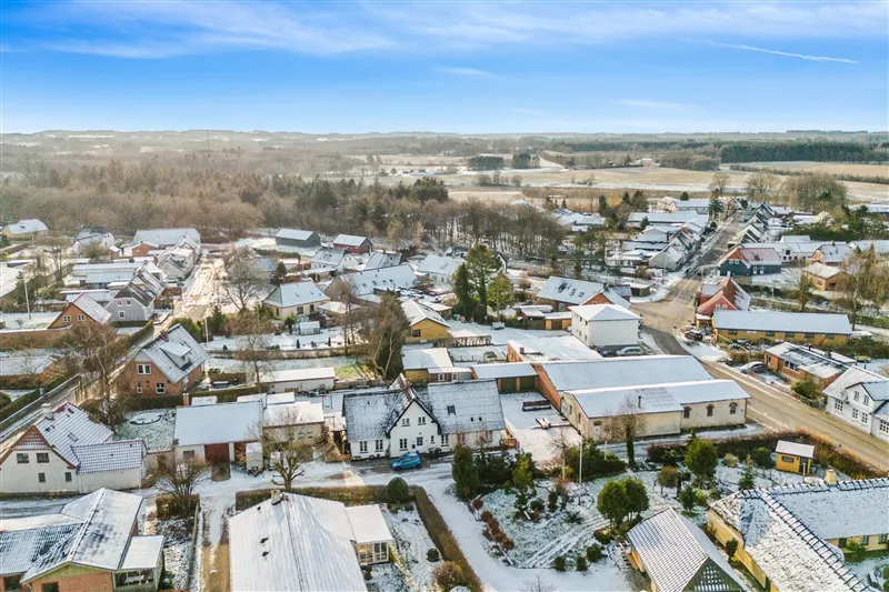 Et lille dansk byområde med snedækkede tag og veje. Huset og garagen er snedækkede, og der er flere biler parkeret.