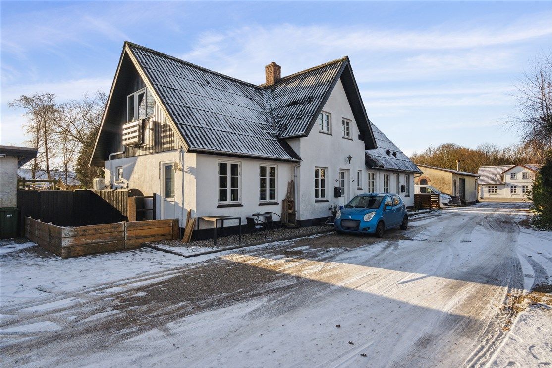 En hvid sommerhusvilla med sort tag i en snepakket parkeringsplads med en blå bil parkeret foran