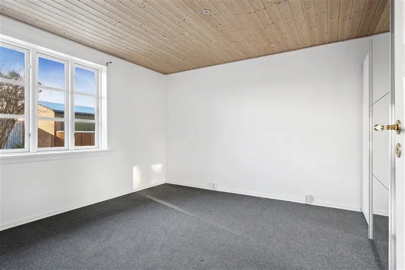 En tom stue med hvide vægge, højt gulv og et vindue med udsigt til en have.