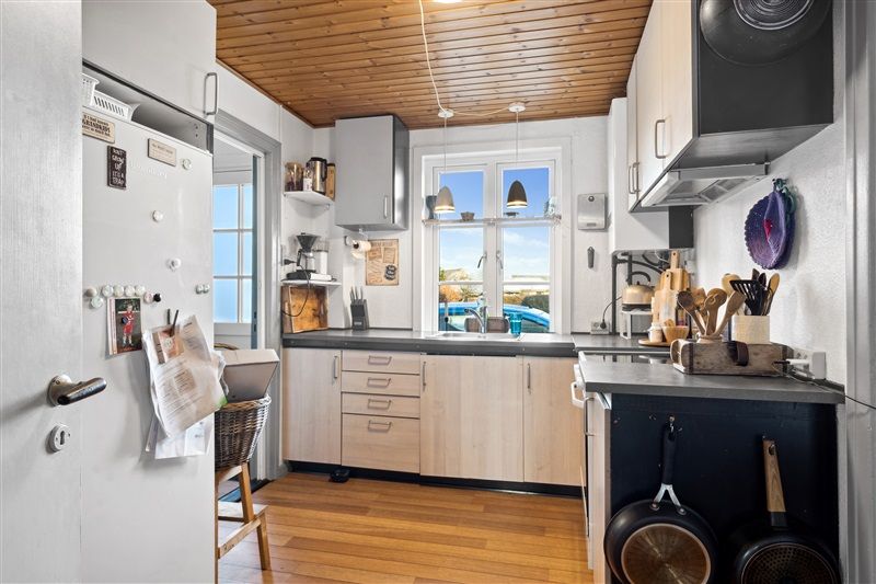 Køkken med lyse lofter, lysegrå vægge og trægulv, udstyret med lyse træskabe og et flot vindue med udsigt til udendørs