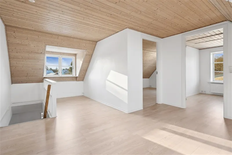 Et lyst værelse med lysegul gulv og hvide vægge samt store vinduer med udsigt til det ydre