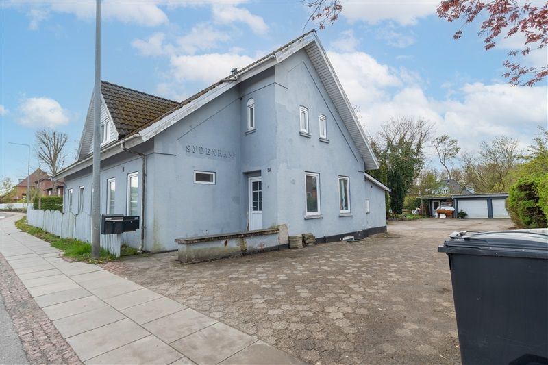 En hvid malet hus med rødt tag og "SYDENHAIM" på facaden, liggende på en landevej med skrapelister og søjler