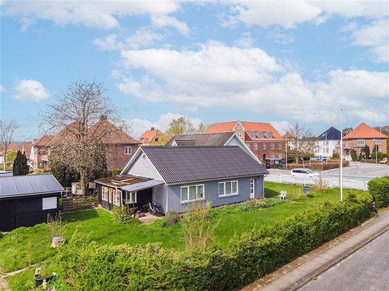 En række huse med røde tag og en bil parkeret på siden af vejen, tæt på en grøn skov og en gårdhave med blomster.