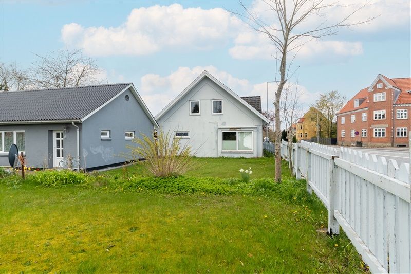 To huse med en grøn have, hvidt panel og højt græs samt en hvid skurtag.