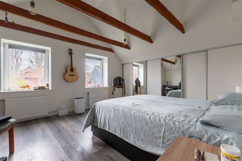 Soveværelse med træloft, to vinduer, akustisk guitar hængende på væggen og et enkelt seng med pudet og spredte tæpper