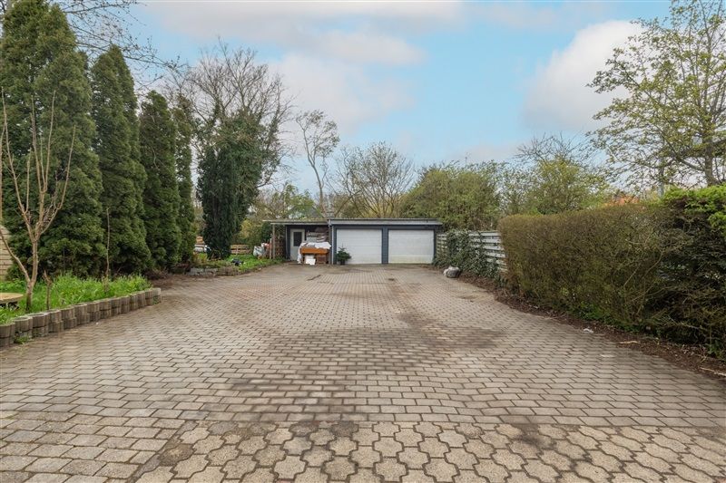 en længstregget vej med brolægning og to garager med hvide døre. Omkring vejen er der grønne buske og træer.