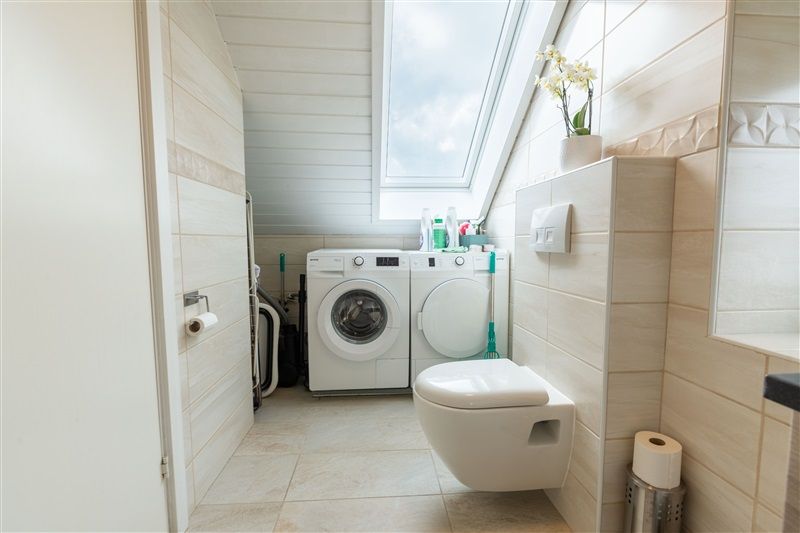 En ren, hvid vaskeri med to maskiner og et toilet samt en lille plante i en gryde