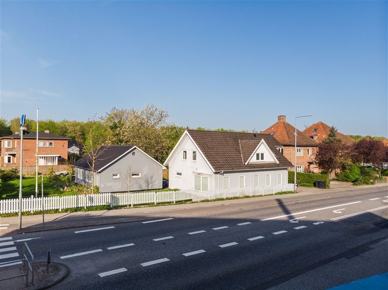 Vej med hvide striber og markeringer, huse med forskellige farver og stilarter, parkeringsmuligheder og grønne anlæg