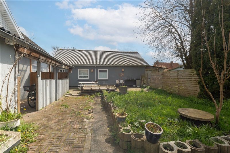 En grøn have med brændenælde, græs, træer og buske, en terrasse med en træbord og stole samt en cykel og blomsterpotter