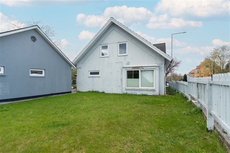 To huse med en grøn græsplæne foran. Huset er hvidt og slankt med lyseruder. Der er et skur og et lysarm bag huset. Der er