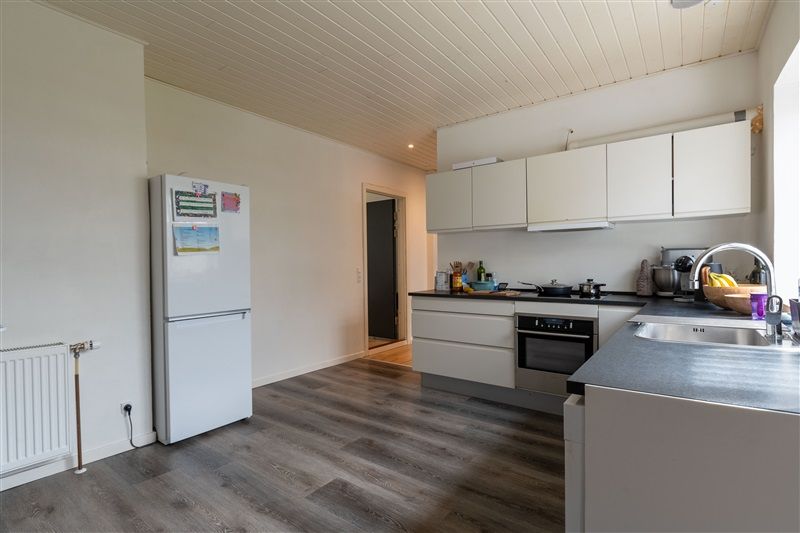 Et moderne køkken med lyse hvide vægge og trægulve samt hvide køkkenskabe og en hvid køleskab.