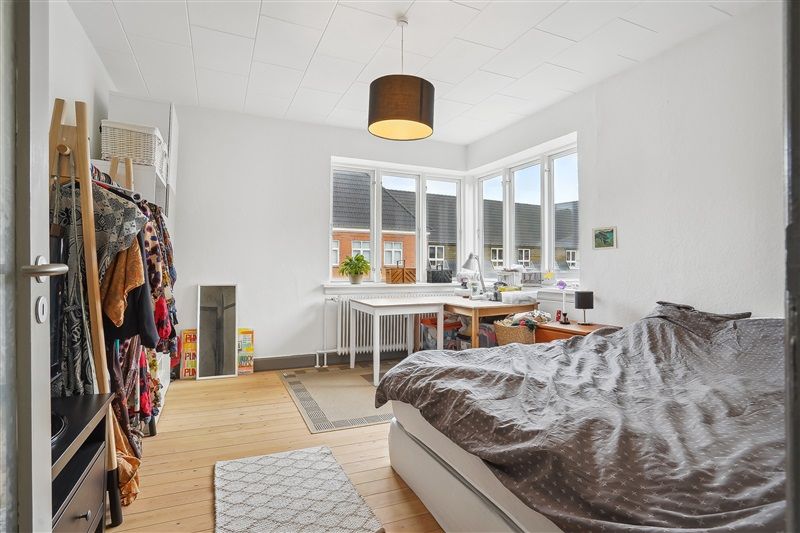 Velholdt soveværelse med hvidt tøjskab, hængende tøj og hvid seng med dyne. På gulvet er der lysegrå tæpper og en bordlampe