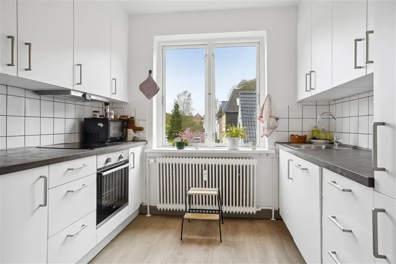 Velholdt køkken med moderne møbler, hvide vægge, køkkenhøj og vinduesvisning med udsigt til træer og bygninger i baggrunden