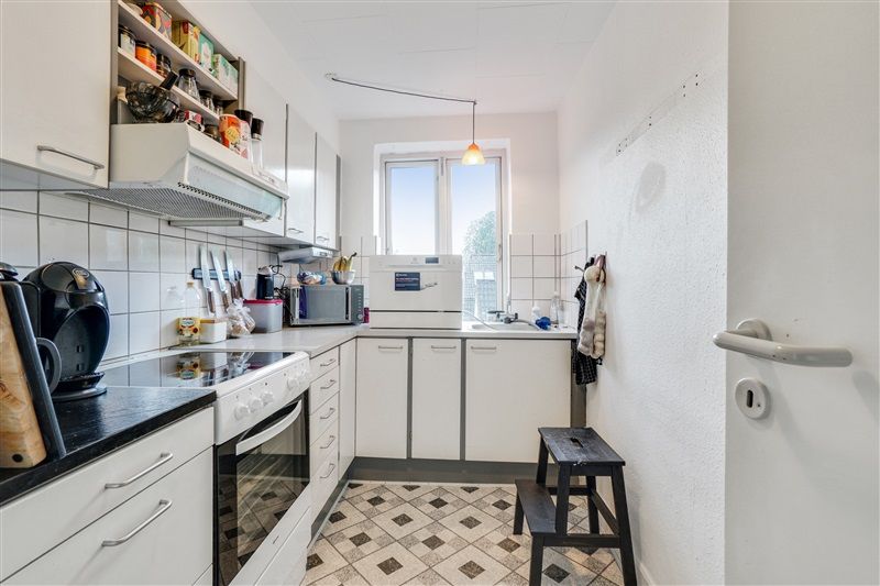 Dansk køkken med hvide vægge, gulve, loft og højre side. Køkken med sort bordplade, sort ovn, hvide loftsskabe, hvide