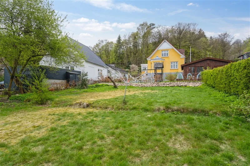 Grønne græspladser med små træer og buske i baggrunden samt hvide og gule husstande med udsigt til skoven