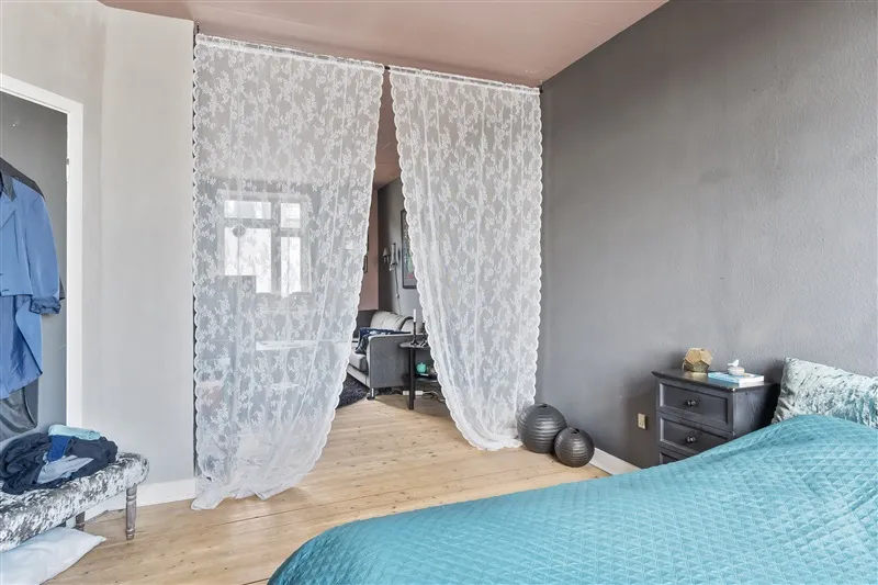 Soveværelse med blå dyne og hvid gardin foran sengen, samt tøj i en stol og en bordlampe i stuen