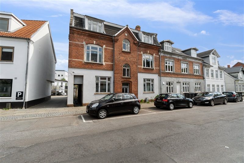 Fire parkerede biler foran et rødt murstenshus og hvide bygninger i baggrunden under et blåt skyfri himmelkuppel