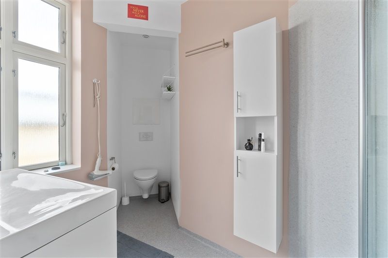 Et badeværelse med hvidt toilet, hvidt badekar og hvide vægge og et højt skab med en toiletbørste og toiletpapir