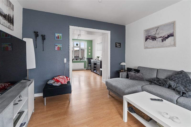 Moderne stue med grå sofa og hvidt bord, hvor fjernsynet står på høj sokkel med hylder, og kunstværk er hængt på væggen