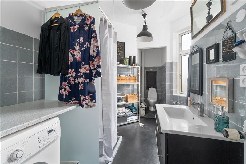 Et lille badeværelse med badekar, håndvask, toilet, håndklædeholder og vasketøjsmaskine samt klædeskab og tøjhængere med tøj