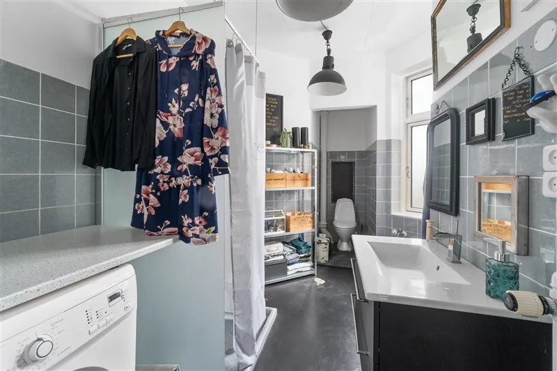 Et lille badeværelse med badekar, håndvask, toilet, håndklædeholder og vasketøjsmaskine samt klædeskab og tøjhængere med tøj