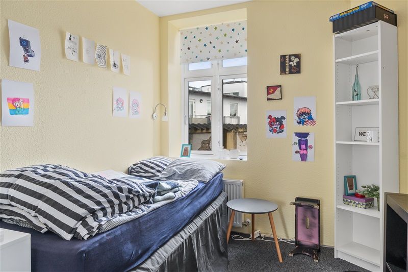 Smukt indrettet værelse med bøf og høj vinduesbue, lysegul væg og lyseblå sengelinned, åben gardin og små billeder