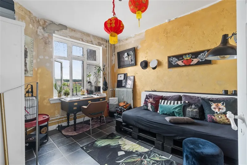 En hyggeligt indrettet stue med gule vægge og sort sofa med flere puder, samt en skrivebord med stole og plante på