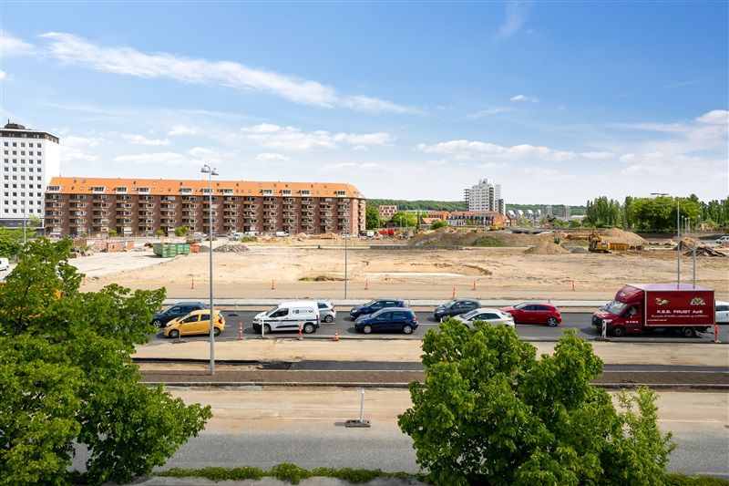 Parkerede biler og lastbiler på en parkeringsplads med bygninger og konstruktion i baggrunden