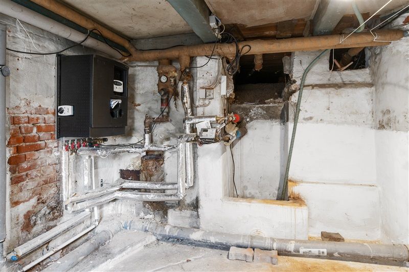 Et gammelt lagerrum med røde mursten og røgskader, gamle rørmærker og knækkede rør samt et sort elektrisk apparat og en