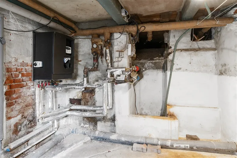 Et gammelt lagerrum med røde mursten og røgskader, gamle rørmærker og knækkede rør samt et sort elektrisk apparat og en