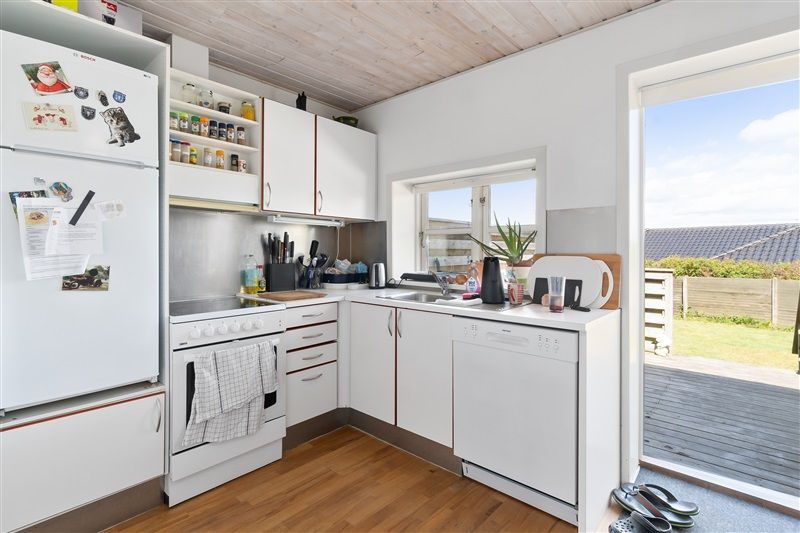 Et moderne køkken med hvide vægge, hvidt køleskab og lysegrå trægulv samt en stor glasdør og udsigt til en grøn have