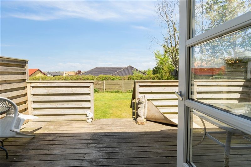 Træterrasse med udsigt til grønt græs og have, et hvidt skur og træhegn med udsigt til skov og hus på afstand