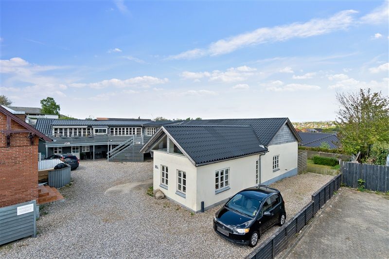 En hvid lejlighed med garage og terrasse ligger i en lejlighedsbygning med parkeringsplads og skur i baggrunden