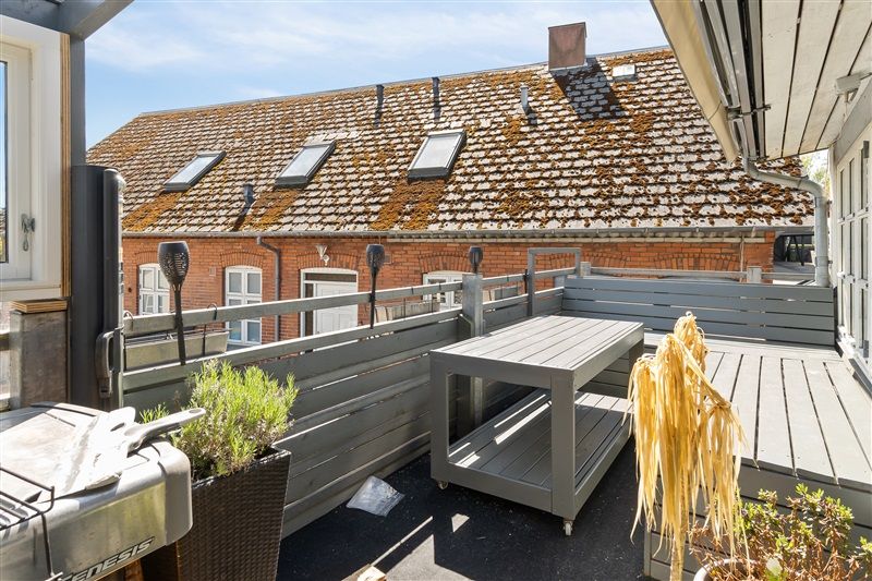 Terrasse med skur, grill, træbord, plante og skurtag. Bagved er et rødt hus med mursten og gule vinduer.