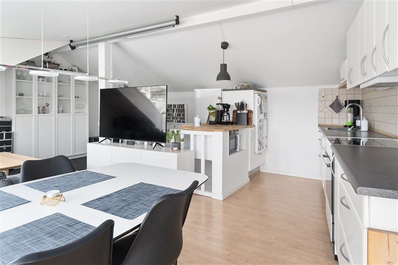 Køkken med lyse vægge og trægulv hvor der er en spiseplads med bord og stole og et køkken med apparater og hængelampe