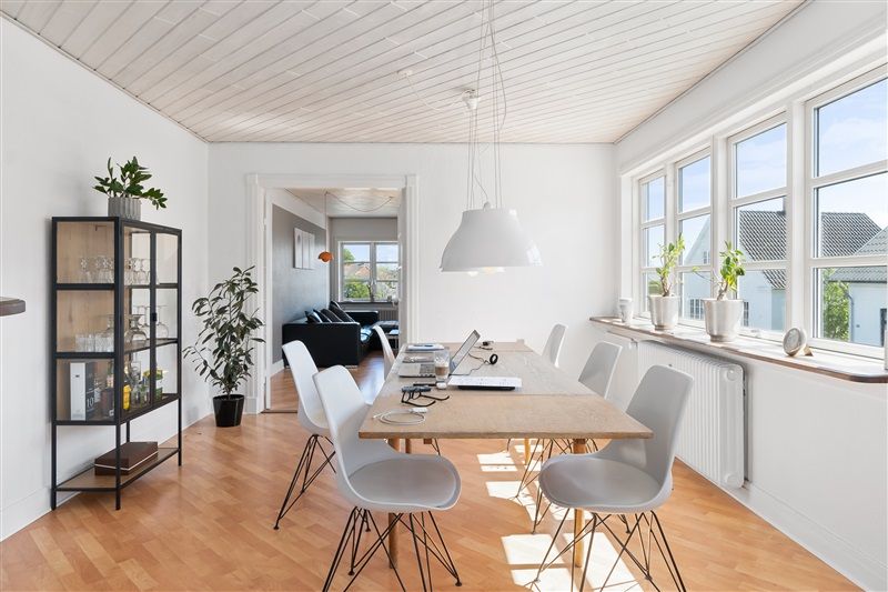 Moderne stue med højt loft, hvide vægge, og lyse trægulve. Bord med flere hvide stole, og en hvid lampe. Planter og glasreol